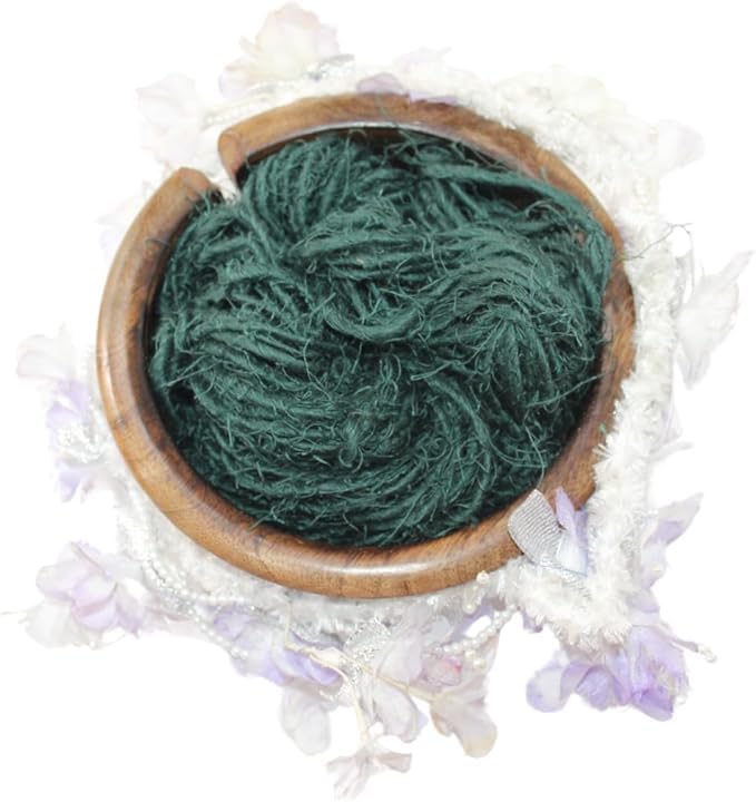 Dark green KnitSilk Artisan linen yarn in a wooden bowl with pale flowers.