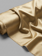 A close-up of a rolled piece of shiny satin fabric in a warm champagne-gold color, draped elegantly against a neutral gray background.